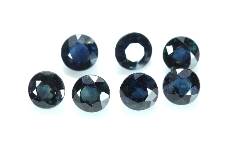 May include: Seven round, faceted, dark blue gemstones. The gemstones are arranged in a semi-circle on a white background.