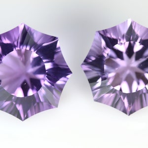 May include: Two faceted, starburst-shaped gemstones in shades of lavender and purple. Each gem has a central circular facet surrounded by pointed, triangular facets. The gems are displayed against a white background.