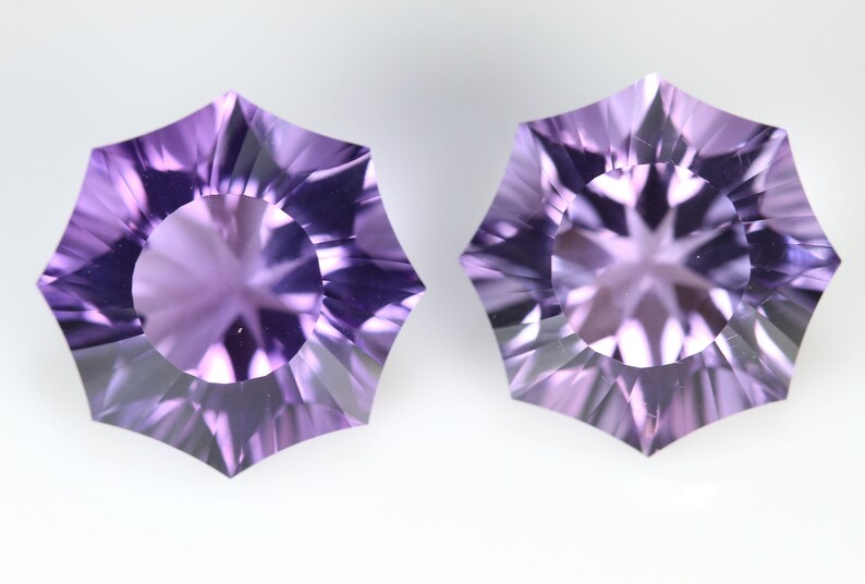 May include: Two faceted, starburst-shaped gemstones in shades of lavender and purple. Each gem has a central circular facet surrounded by pointed, triangular facets. The gems are displayed against a white background.