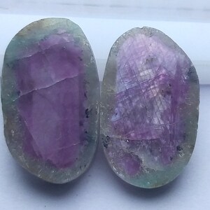 May include: Two polished, oval-shaped gemstones. The stones have a vibrant purple centre with a green and grey border. The surfaces display natural patterns and textures, suggesting a raw, organic origin. The gemstones are displayed on a white background.