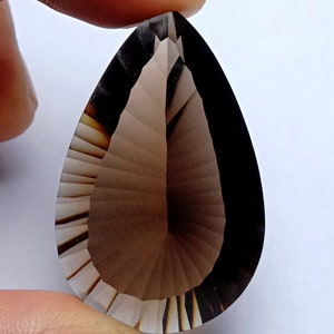 May include: A faceted, teardrop-shaped gemstone with a gradient of brown hues. The gem is held between fingers, showcasing its intricate cuts and polished surface. The stone's color transitions from light to dark, highlighting its clarity and form.