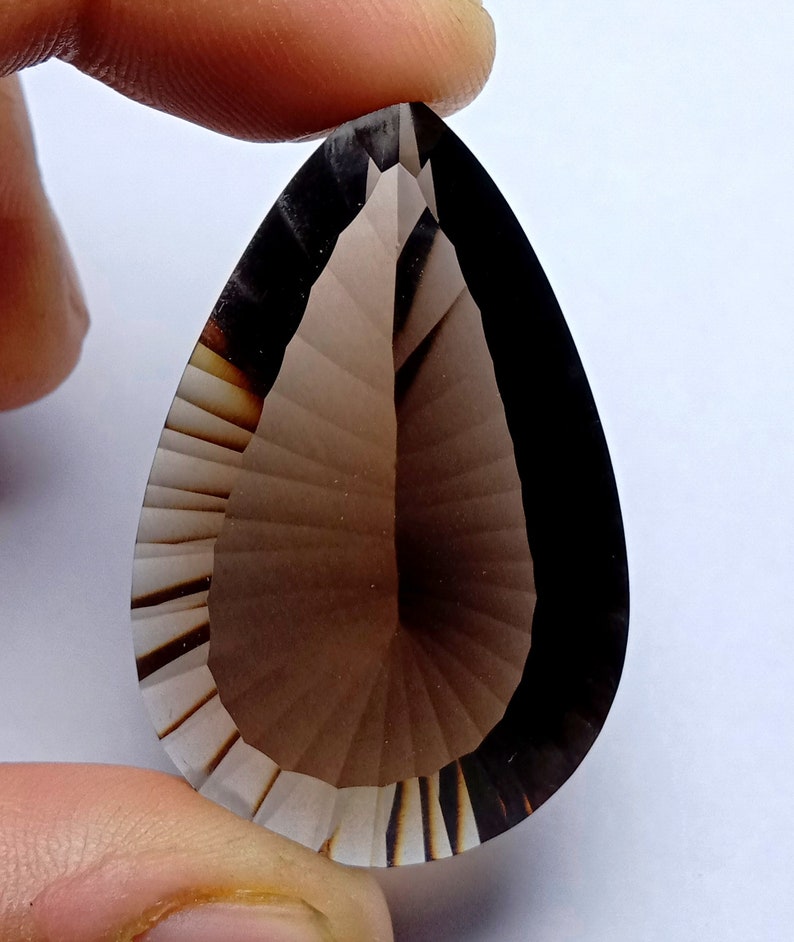 May include: A faceted, teardrop-shaped gemstone with a gradient of brown hues. The gem is held between fingers, showcasing its intricate cuts and polished surface. The stone's color transitions from light to dark, highlighting its clarity and form.