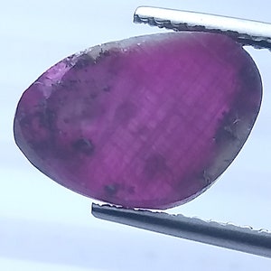 May include: A polished, oval gemstone in deep magenta and purple hues, held by silver tweezers. The gem has a smooth surface with subtle colour variations and small dark inclusions. The overall shape is slightly irregular.