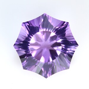May include: A faceted, star-shaped gemstone in shades of purple. The gem has a complex, multi-faceted design that reflects light, creating a sparkling effect. The gemstone is set against a white background.