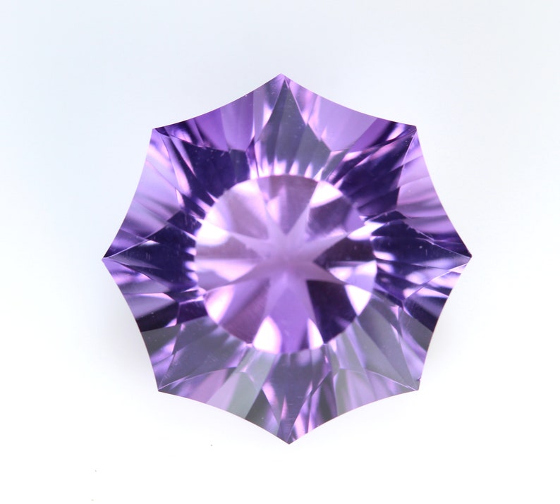 May include: A faceted, star-shaped gemstone in shades of purple. The gem has a complex, multi-faceted design that reflects light, creating a sparkling effect. The gemstone is set against a white background.