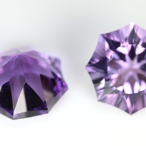 May include: Two faceted, starburst-cut gemstones in shades of purple. The gems have a symmetrical, multi-faceted design, with a central point and radiating facets. The gemstones are set against a white background, highlighting their color and cut.