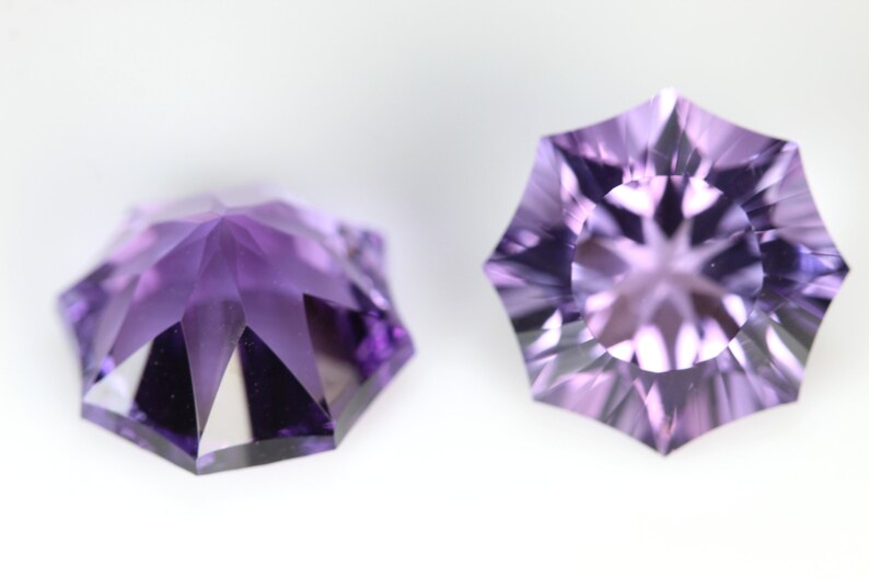 May include: Two faceted, starburst-cut gemstones in shades of purple. The gems have a symmetrical, multi-faceted design, with a central point and radiating facets. The gemstones are set against a white background, highlighting their color and cut.