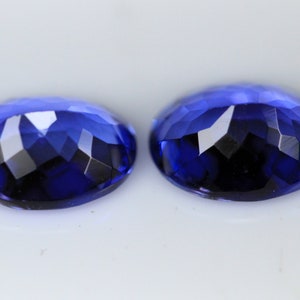 May include: Two faceted, oval-shaped gemstones in a deep, rich blue hue. The gems have a brilliant cut, reflecting light with multiple facets. The stones are set against a white background, highlighting their color and clarity.