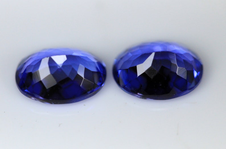 May include: Two faceted, oval-shaped gemstones in a deep, rich blue hue. The gems have a brilliant cut, reflecting light with multiple facets. The stones are set against a white background, highlighting their color and clarity.