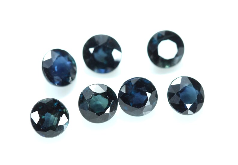 May include: Seven oval-shaped, dark blue gemstones with a faceted cut. The gemstones are arranged in a scattered pattern on a white background.
