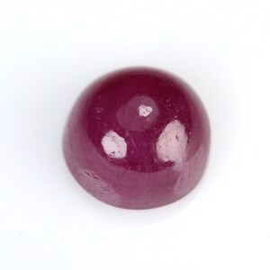 May include: A cabochon-cut ruby gemstone, a deep, rich red color, is displayed against a white background. The smooth, rounded surface of the gemstone reflects light, showcasing its polished finish. This gemstone is suitable for jewelry making.