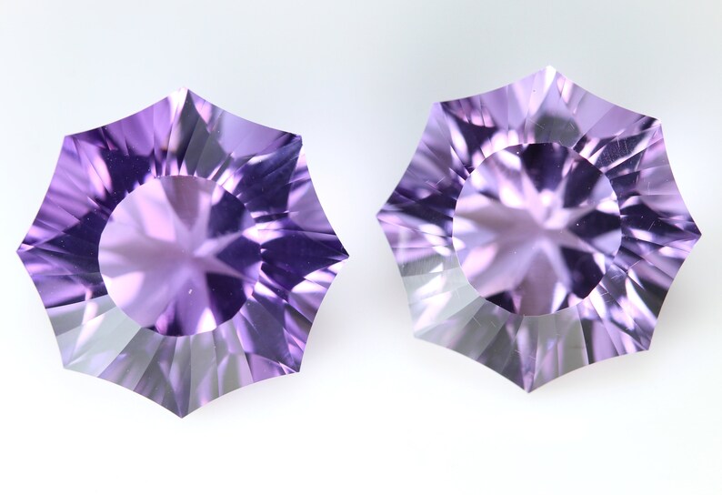 May include: Two faceted, starburst-shaped gemstones with a light purple hue. The gems have a central circular facet surrounded by radiating, triangular facets. The background is plain white, highlighting the gems' intricate cuts and color.