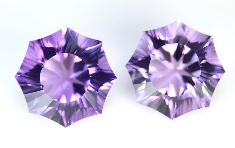 May include: Two faceted, light purple gemstones with a starburst pattern in the center. The gems have a unique, multi-sided shape, and are set against a white background. These gemstones are ideal for jewelry making.