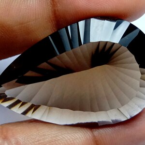 May include: A pear-shaped, faceted gemstone with a smoky gray hue. The gem features a detailed, fan-like pattern carved into its surface, creating a play of light and shadow. The stone is held by a hand, showcasing its size and shape.