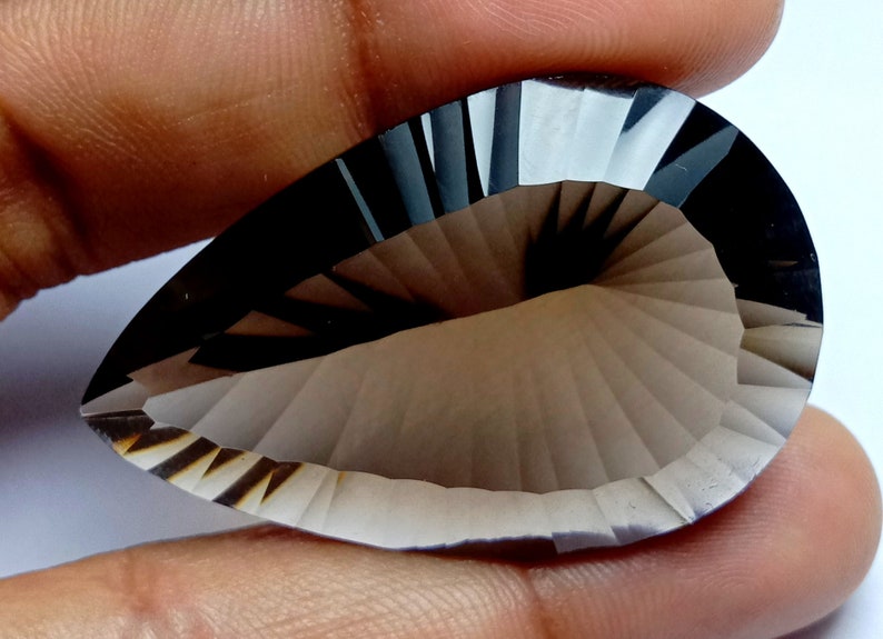 May include: A pear-shaped, faceted gemstone with a smoky gray hue. The gem features a detailed, fan-like pattern carved into its surface, creating a play of light and shadow. The stone is held by a hand, showcasing its size and shape.