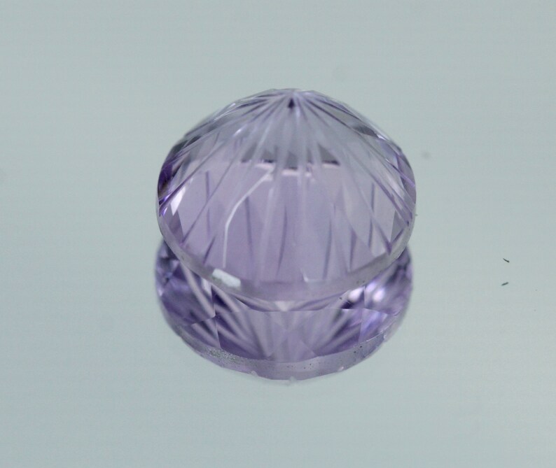 May include: A faceted amethyst gemstone with a purple hue. The gemstone is cut into a round shape with a pointed top.
