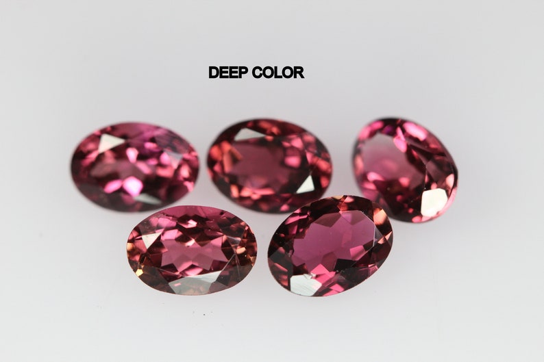 May include: Five oval-shaped gemstones with a deep pink color. The gemstones are arranged in a semi-circle on a white surface. The text "DEEP COLOR" is written above the gemstones.