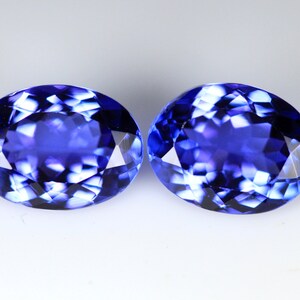 May include: Two oval-cut, faceted gemstones with a deep blue color. The gems have a brilliant, reflective surface, showcasing internal patterns. The stones are set against a plain white background, highlighting their color and cut.