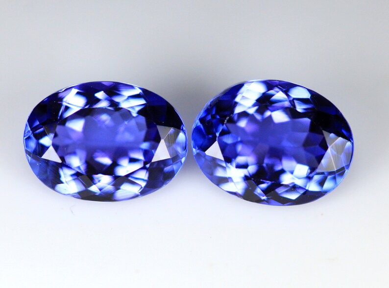 May include: Two oval-cut, faceted gemstones with a deep blue color. The gems have a brilliant, reflective surface, showcasing internal patterns. The stones are set against a plain white background, highlighting their color and cut.