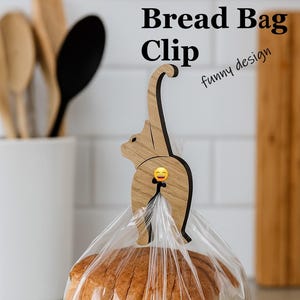 May include: A wooden bread bag clip in the shape of a cat with a smiling emoji. The clip is holding a clear plastic bag of sliced bread. Text on the image reads "Bread Bag Clip" and "funny design".