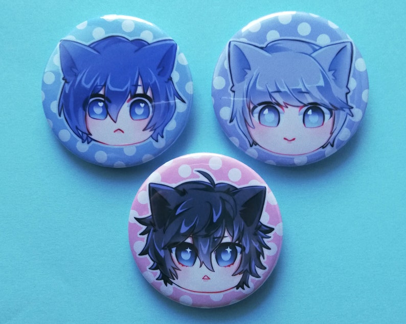May include: Three round buttons featuring cartoon characters. Each button has a different coloured background: blue, pink, and light blue. Each character has cat ears and blue eyes.
