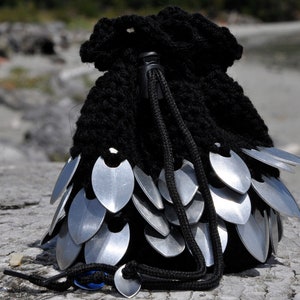 May include: Black crocheted drawstring bag with silver metal scales. The bag has a black drawstring and is closed.