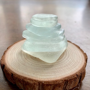 May include: A stack of smooth, light blue sea glass pieces arranged in a pyramid shape on a wooden slice. The glass is translucent and has a slightly frosted appearance.