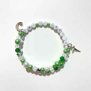 May include: A beaded bracelet with white letter beads spelling "AND TO SATURN". The bracelet features green and white beads, silver star charms, and a crescent moon charm. The bracelet is a celestial-themed accessory.