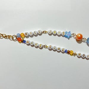 Peut inclure: Un porte-clés doré avec deux brins perlés. Le brin supérieur épelle "KILL JOYS" avec des perles de lettres blanches, une perle étoile bleue et des perles orange et bleues. Le brin inférieur dit "MAKE SOME NOISE" avec des perles de lettres blanches et des perles colorées.