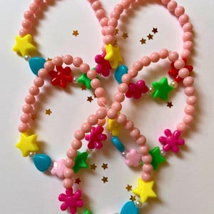 Può includere: Quattro braccialetti di perline rosa con ciondoli a forma di stella e fiore colorati. I braccialetti sono disposti in cerchio su uno sfondo bianco con glitter dorati.