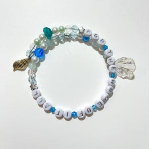 May include: A handmade bracelet with white letter beads spelling "FOR THE LOVE OF IT". The bracelet features blue and clear beads, pearls, a gold seashell charm, and a clear shell charm. The bracelet is a unique piece of jewellery.