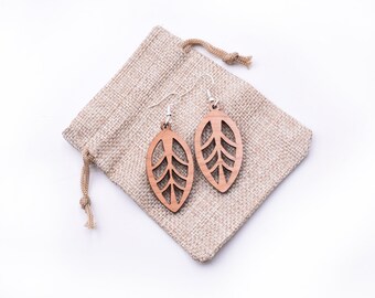 Puede incluir: Un par de pendientes de madera con forma de hoja y un delicado diseño de corte. Los pendientes se muestran en una pequeña bolsa de arpillera.