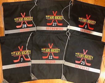 May include: Six black drawstring bags with a white reflective stripe and a yellow and red hockey stick graphic. The bags are personalized with the text "Titans Hockey" and a player's name and number.