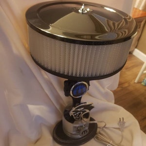 May include: A black and silver table lamp with a chrome air filter shade and a Mopar logo. The lamp base features a silver car part with a black and silver Mopar logo.