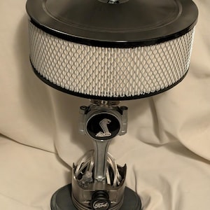 May include: A unique lamp crafted from automotive parts. It features a black air filter, a silver connecting rod with a Cobra emblem, and a piston base with a Ford logo. The lamp's design incorporates industrial and automotive elements.