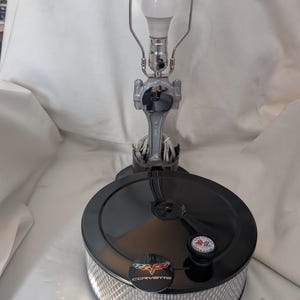 May include: A unique lamp crafted from automotive parts, featuring a black air filter base with a Corvette logo and a silver metal rod. The lamp includes a light bulb and a silver lamp frame.