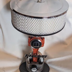 May include: A custom-made lamp featuring a chrome air filter as the lampshade, supported by a red and silver engine piston and connecting rod. The base is a black circular platform, with the MOPAR and HEMI logos visible.