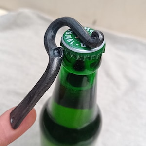 Pode incluir: Um abridor de garrafas de metal escuro forjado à mão é posicionado sobre uma garrafa de vidro verde. O abridor tem uma alça curva e um gancho para levantar a tampa da garrafa. A tampa da garrafa é verde e prateada.