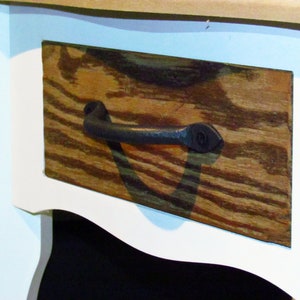 Könnte beinhalten: Nahaufnahme einer Holzschubladenfront mit einem dunklen, gebogenen Metallgriff. Die Holzmaserung ist sichtbar, und die Schublade ist Teil eines weiß lackierten Möbelstücks. Der Griff ist im Stil von dunklem Schmiedeeisen.