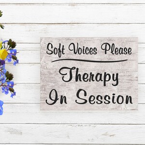 May include: A white wooden sign with black text that reads "Soft Voices Please Therapy In Session". The sign is on a white wood background with a bouquet of colorful flowers to the left.