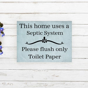 May include: A blue sign with white text that reads "This home uses a Septic System. Please flush only Toilet Paper." The sign is on a white wood background.