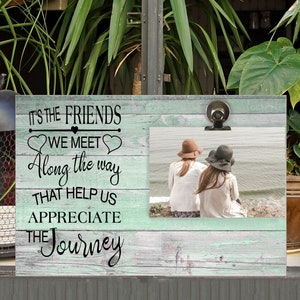 May include: A light blue wooden picture frame with a white clip. The frame has a quote that reads "It's the friends we meet along the way that help us appreciate the journey." The frame has a photo of two people sitting on a beach.