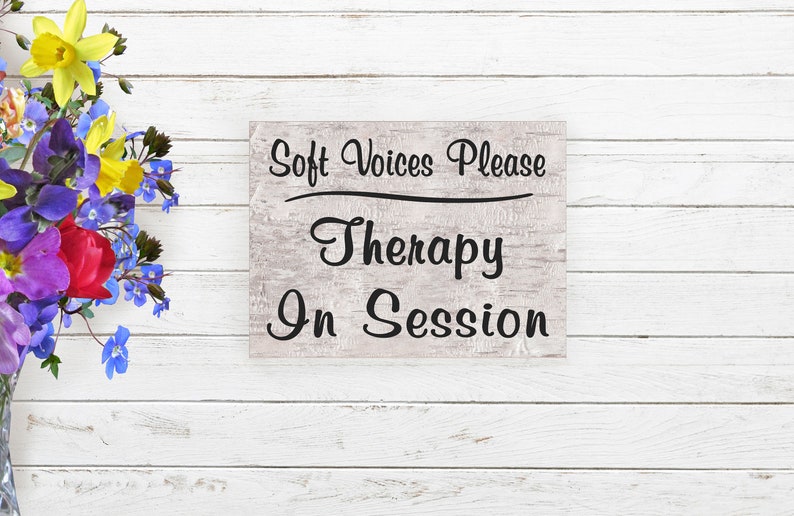 May include: A white wooden sign with black text that reads "Soft Voices Please Therapy In Session". The sign is on a white wood background with a bouquet of colorful flowers to the left.