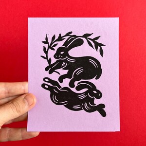May include: A black and white linocut print of two rabbits on a light purple background. The rabbits are in a playful pose, with one rabbit jumping over the other. The print is framed by a simple border.