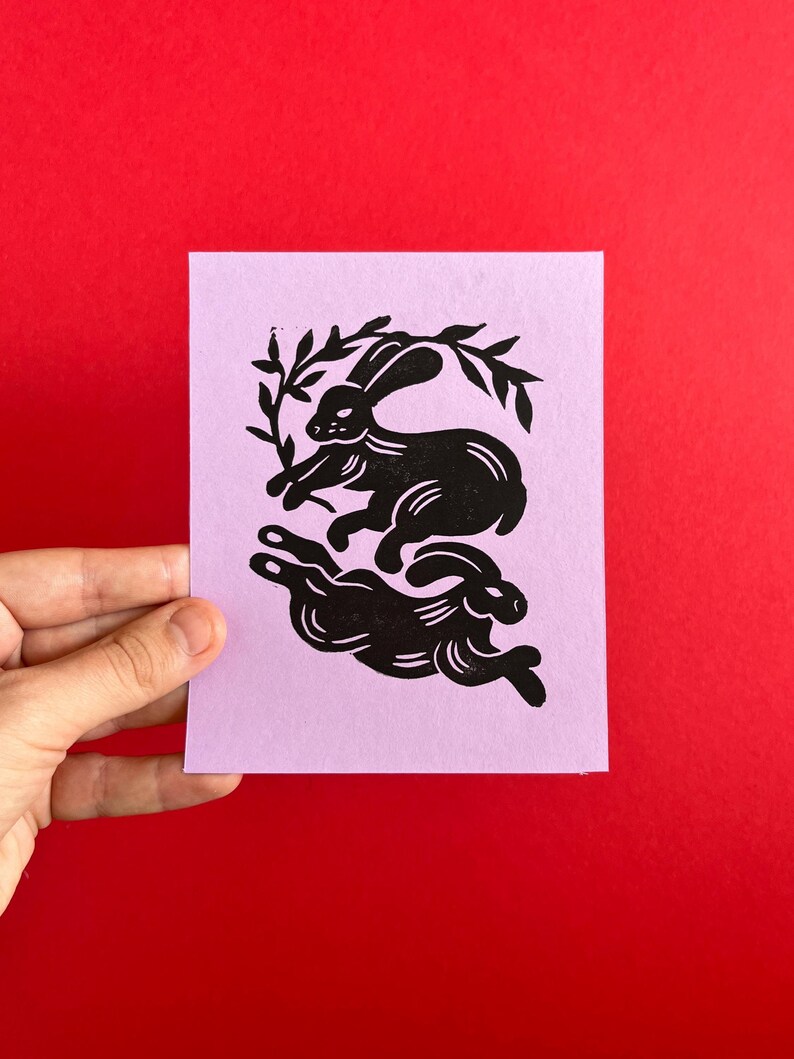 May include: A black and white linocut print of two rabbits on a light purple background. The rabbits are in a playful pose, with one rabbit jumping over the other. The print is framed by a simple border.