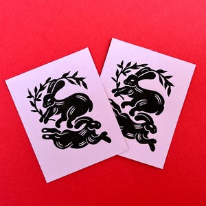 May include: Two pink cards with black ink illustrations of rabbits. The rabbits are in a circular design with leaves and branches.