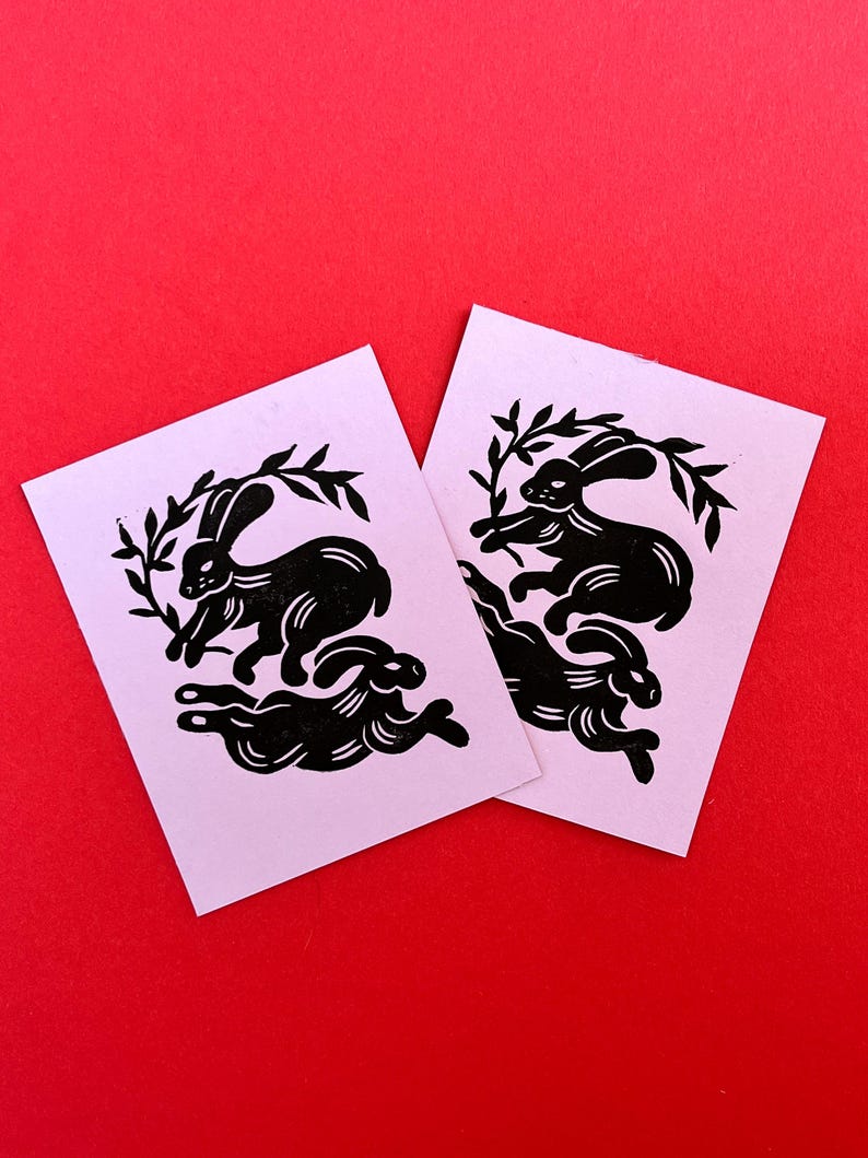 May include: Two pink cards with black ink illustrations of rabbits. The rabbits are in a circular design with leaves and branches.