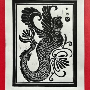 May include: Black and white linocut print of a mermaid with wings and a crown. The mermaid is surrounded by stylized floral designs.
