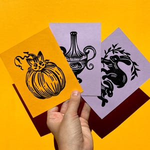 May include: Three black and white linocut prints on different colored paper. The first print features a cat inside a pumpkin, the second print features a vase with a floral design, and the third print features a rabbit with a leafy branch.