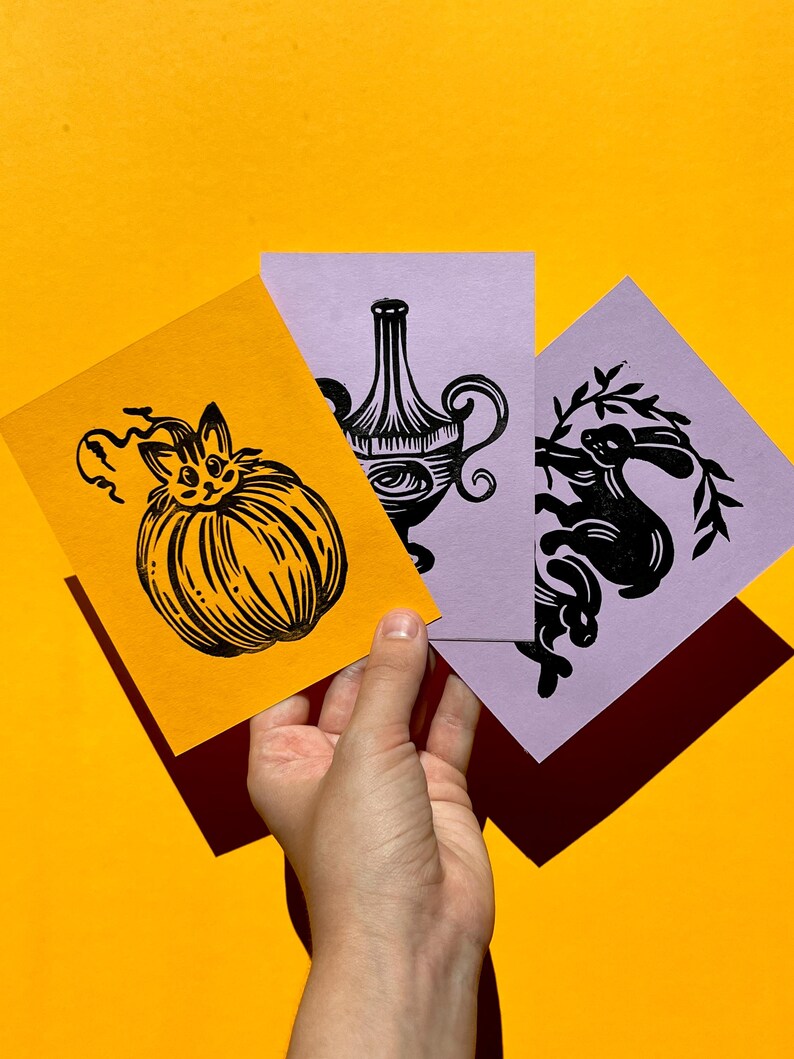 May include: Three black and white linocut prints on different colored paper. The first print features a cat inside a pumpkin, the second print features a vase with a floral design, and the third print features a rabbit with a leafy branch.